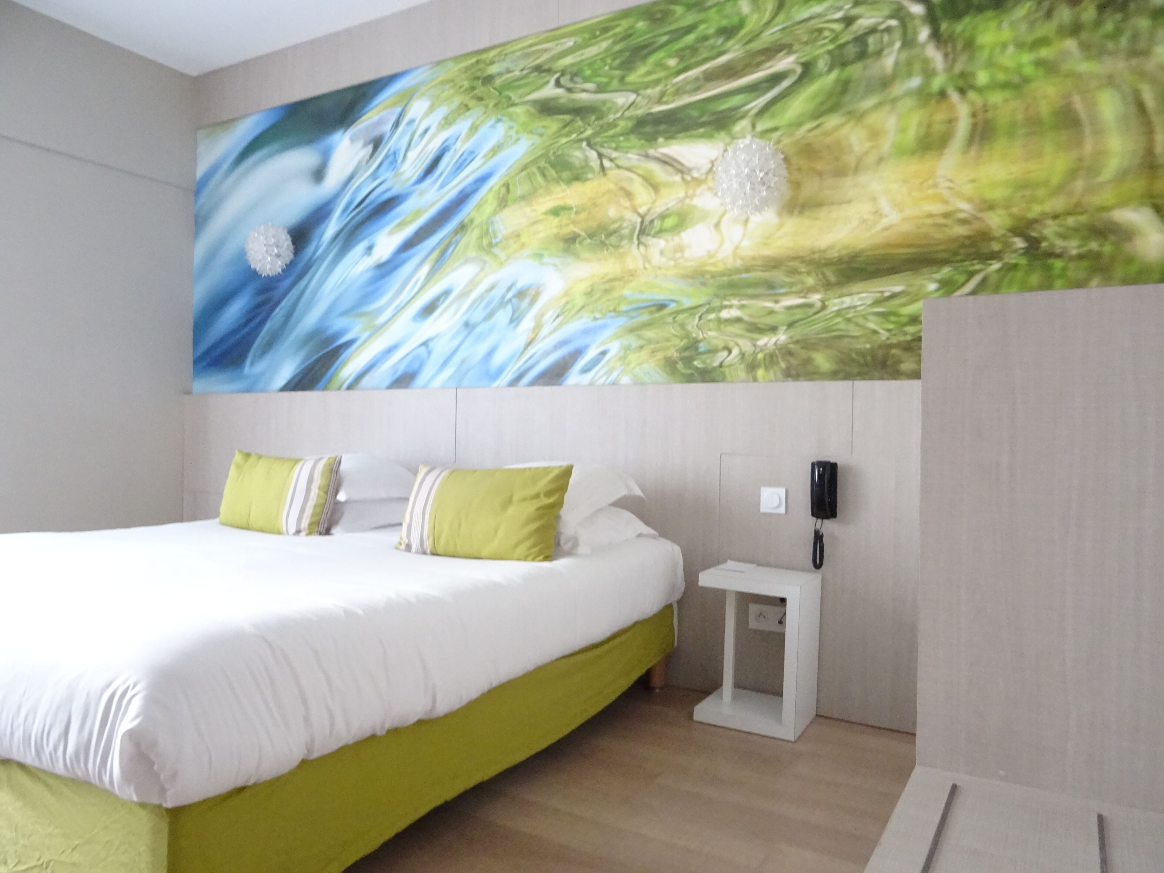 Quarto de hotel moderno com uma cama grande, roupa de cama verde e branca, uma mesa de cabeceira com telefone e um quadro abstrato grande na parede.
