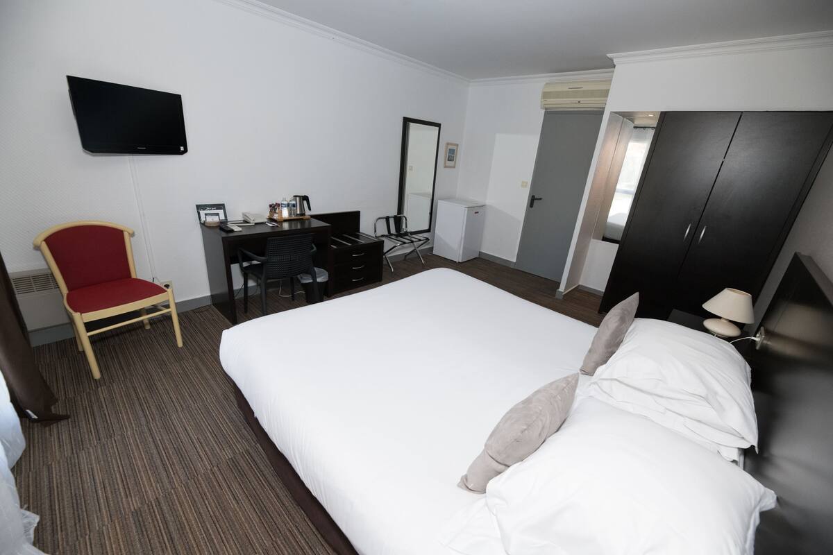 Ein Hotelzimmer mit einem großen Bett, einem Schreibtisch mit Computer, einem Sessel, einem an der Wand befestigten Fernseher und einem Kleiderschrank.