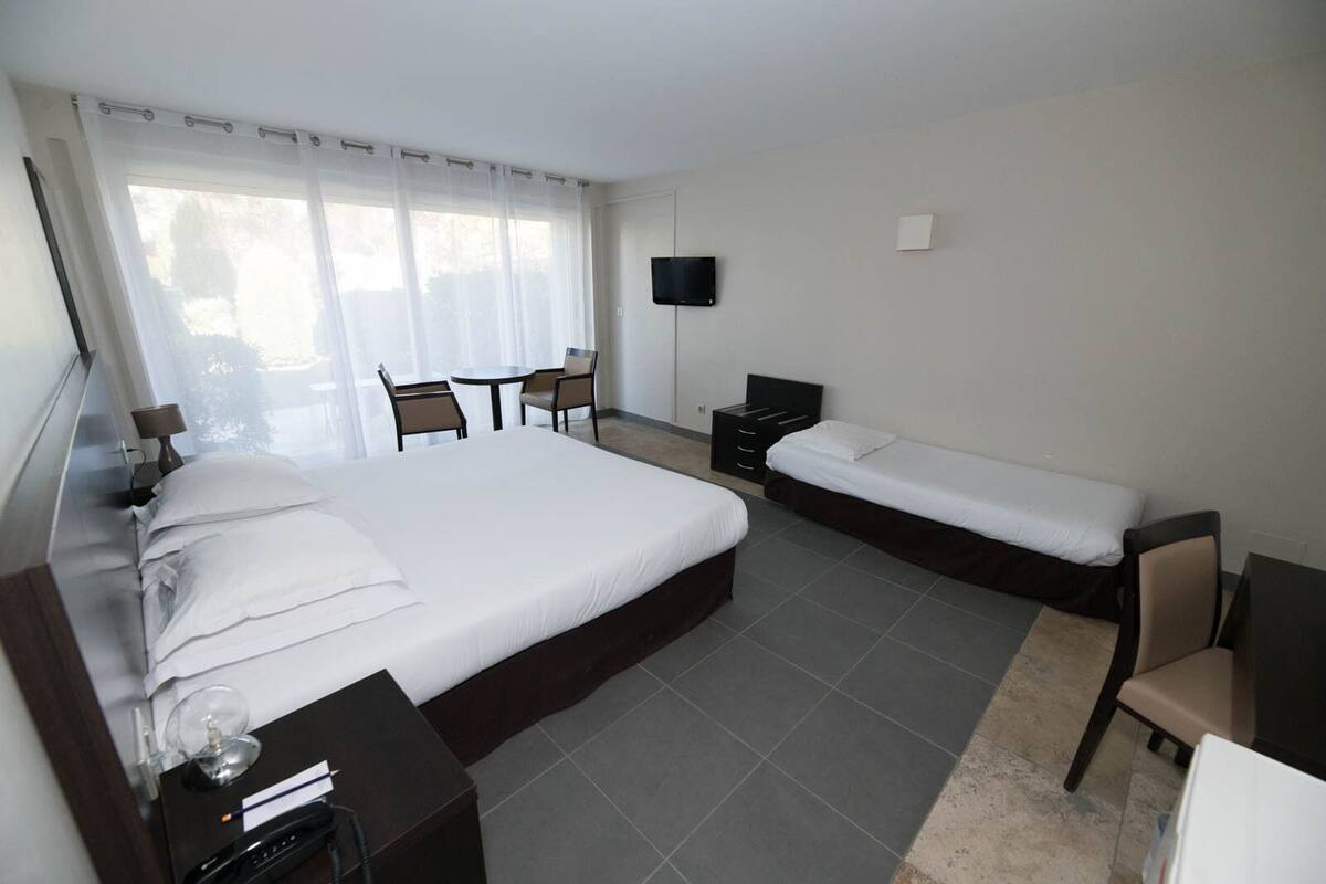 Ein Hotelzimmer mit einem großen Bett, einem kleineren Bett, einem Schreibtisch, einem Sessel, einem Fernseher und einem Fenster mit Vorhängen.