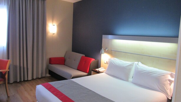 Une chambre d’hôtel avec un grand lit, un canapé et un fauteuil. On y trouve une table de chevet avec un téléphone et une lampe. La chambre a un mur de couleur sombre et une fenêtre avec des rideaux.