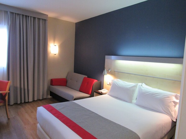 A hotel room with a large bed, a sofa, and a chair. There is a nightstand with a phone and a lamp. The room has a dark wall and a window with curtains. - Holiday Inn Express Madrid-Alcobendas by IHG (Alcobendas)