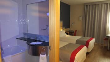 Habitación de hotel con bañera, lavamanos, espejo, escritorio, dos camas y ventana con cortinas.