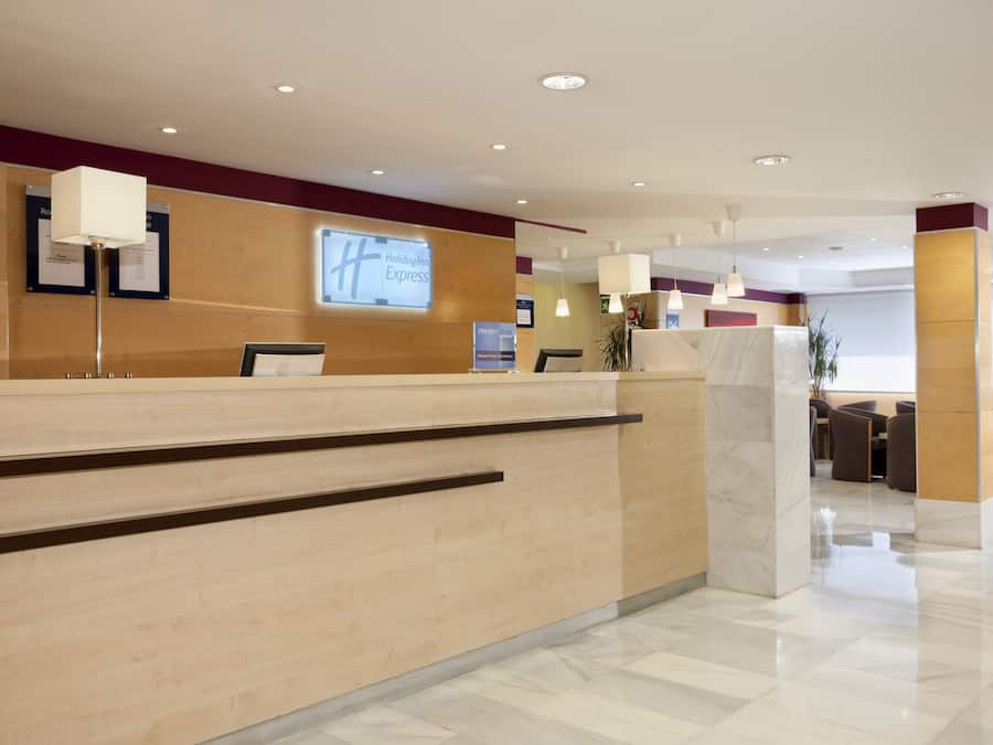 A clean, modern hotel reception area with a wooden reception desk, a 'Holiday Inn Express' sign, and a 'Priority Club' sign.