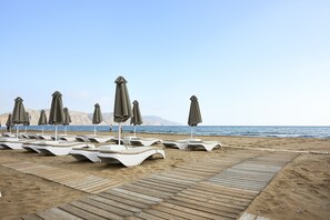 A beach with rows of sun loungers and umbrellas, a wooden walkway, and a view of the sea and mountains. - Mythos Palace Resort & Spa - All Inclusive (Apokoronas)