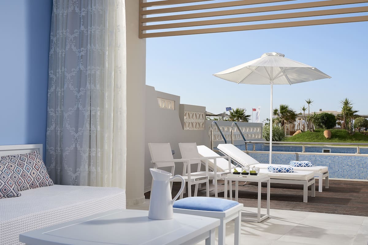 A hotel room with a bed, a white table with a pitcher, and a view of a pool area with lounge chairs and an umbrella.