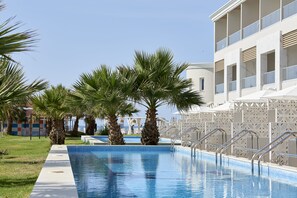 En swimmingpool omgiven av palmer, en vit byggnad med balkonger och en klarblå himmel.