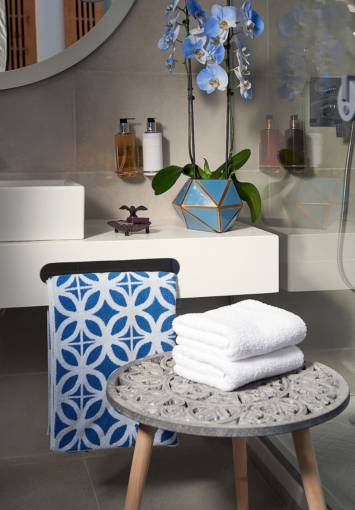 A bathroom with a white bathtub, a blue patterned chair, a round table with white towels, and blue orchids in a geometric vase.