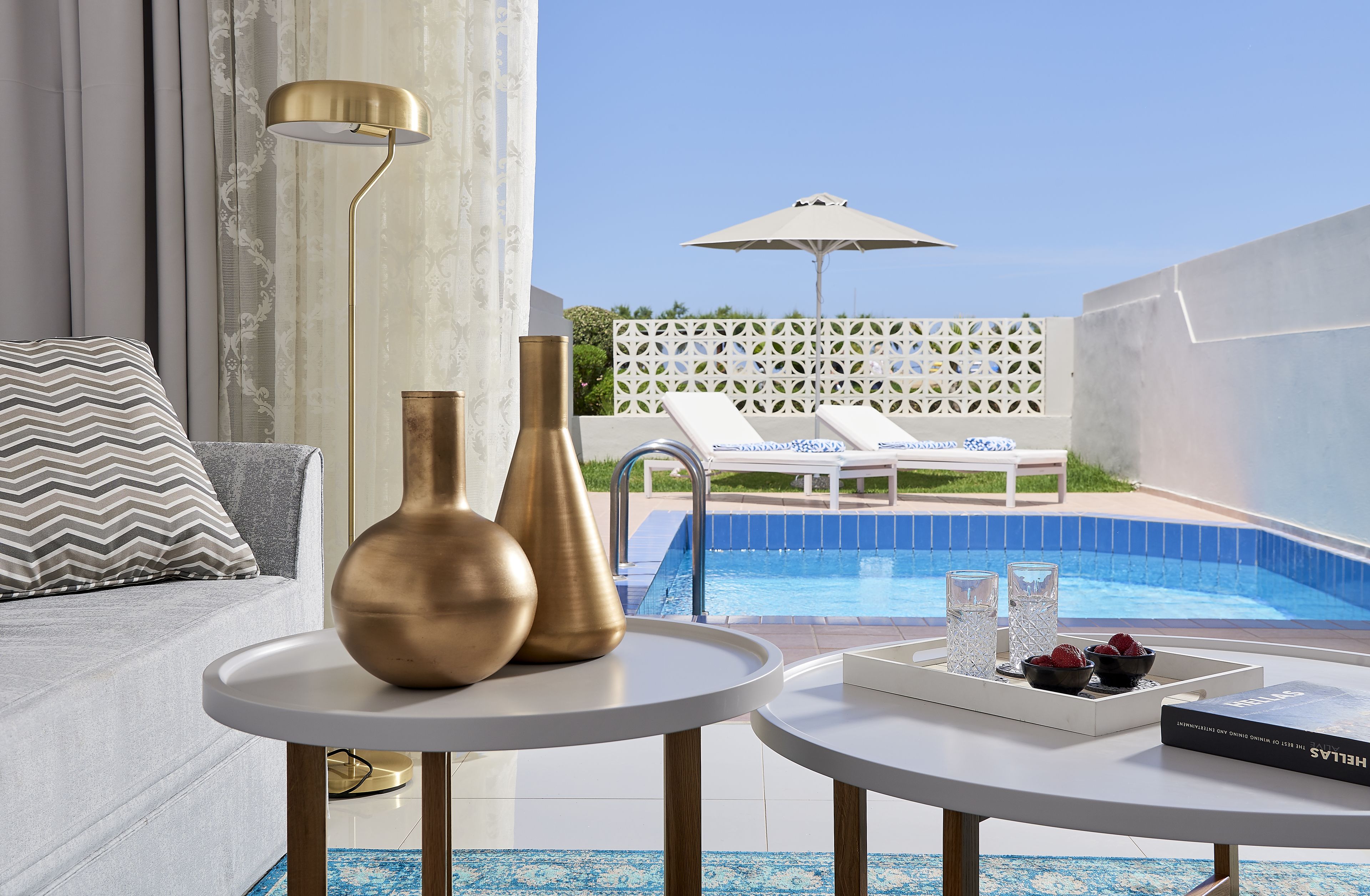 Una camera moderna con vista piscina, con un divano, due tavolini con vasi decorativi, poltrone e un libro.