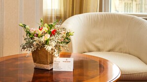 A round wooden table with a floral arrangement and a name card. A beige sofa is in the background with a view of a building through a window. - Boutique Splendid Hotel (Varna)