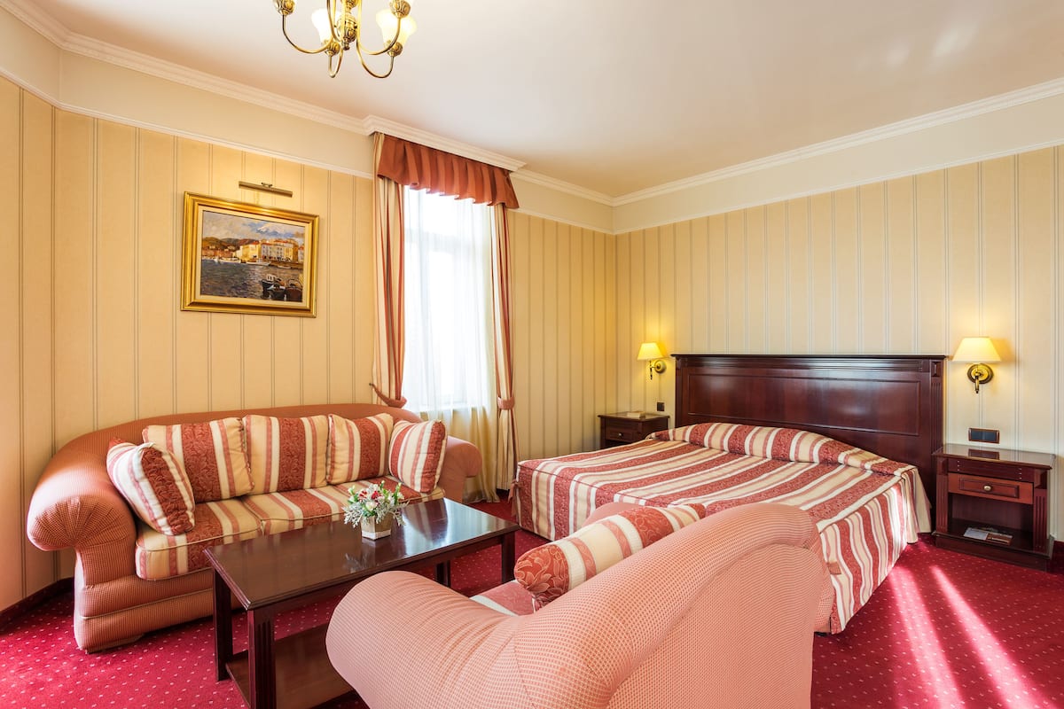 A hotel room with a sofa, two single beds, a wooden nightstand, a table, and a painting on the wall.