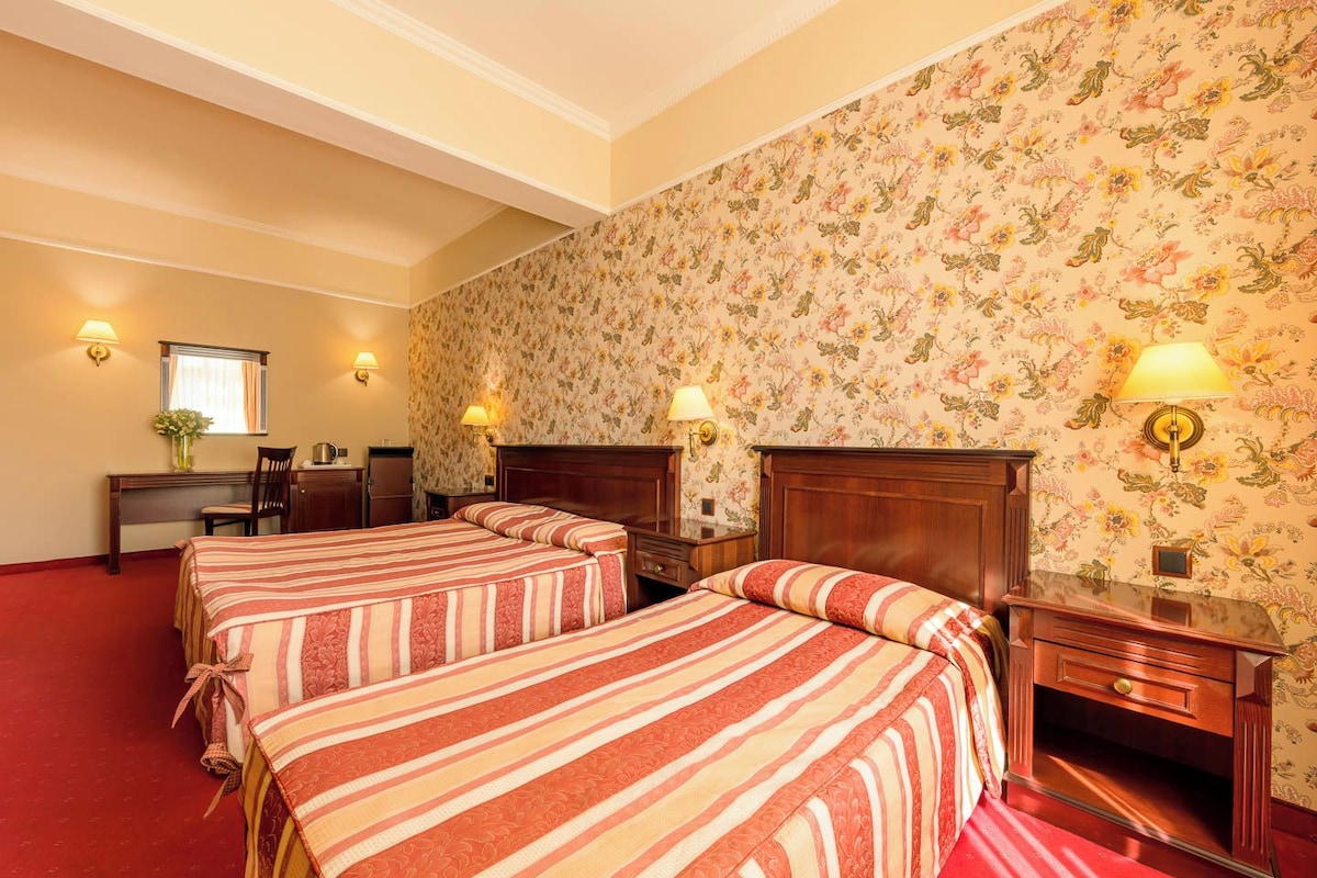 A hotel room with two beds, a desk, and a chair. There are wall sconces and a mirror.