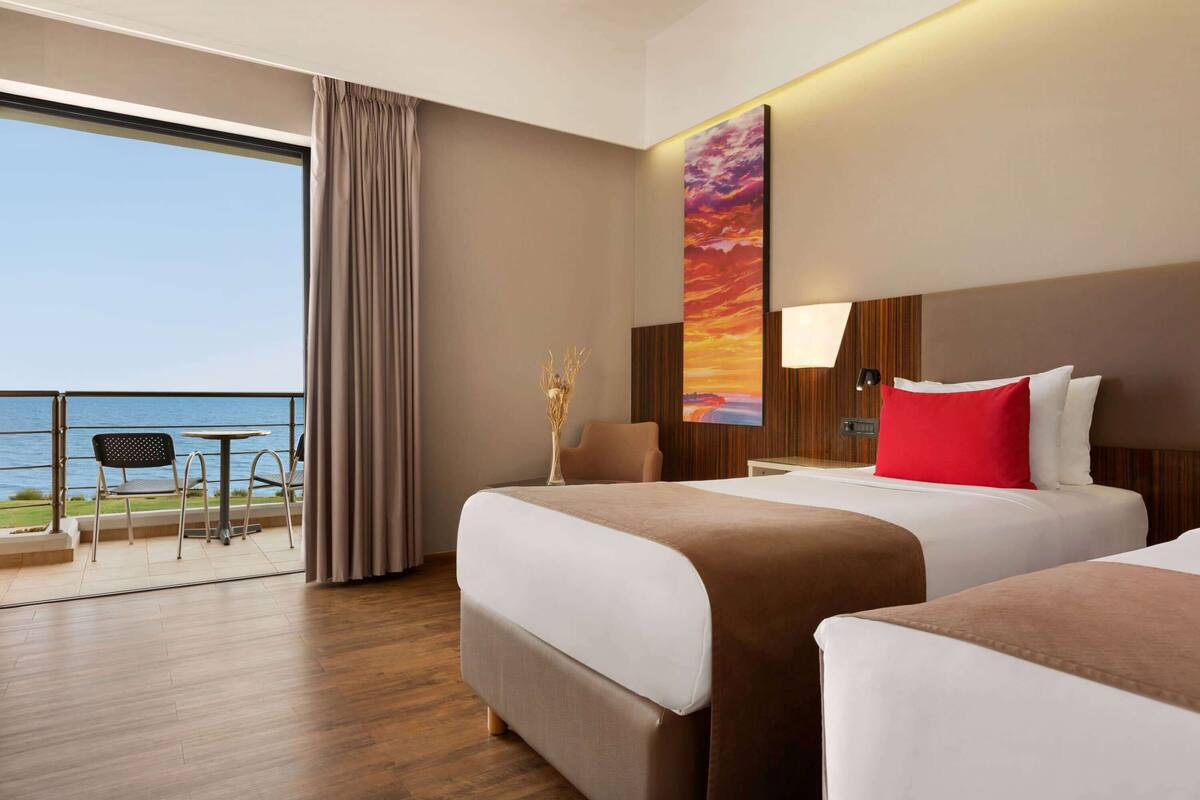 A hotel room with two beds, a large window with a view of the sea, a table and chairs outside, and a sunset painting on the wall.
