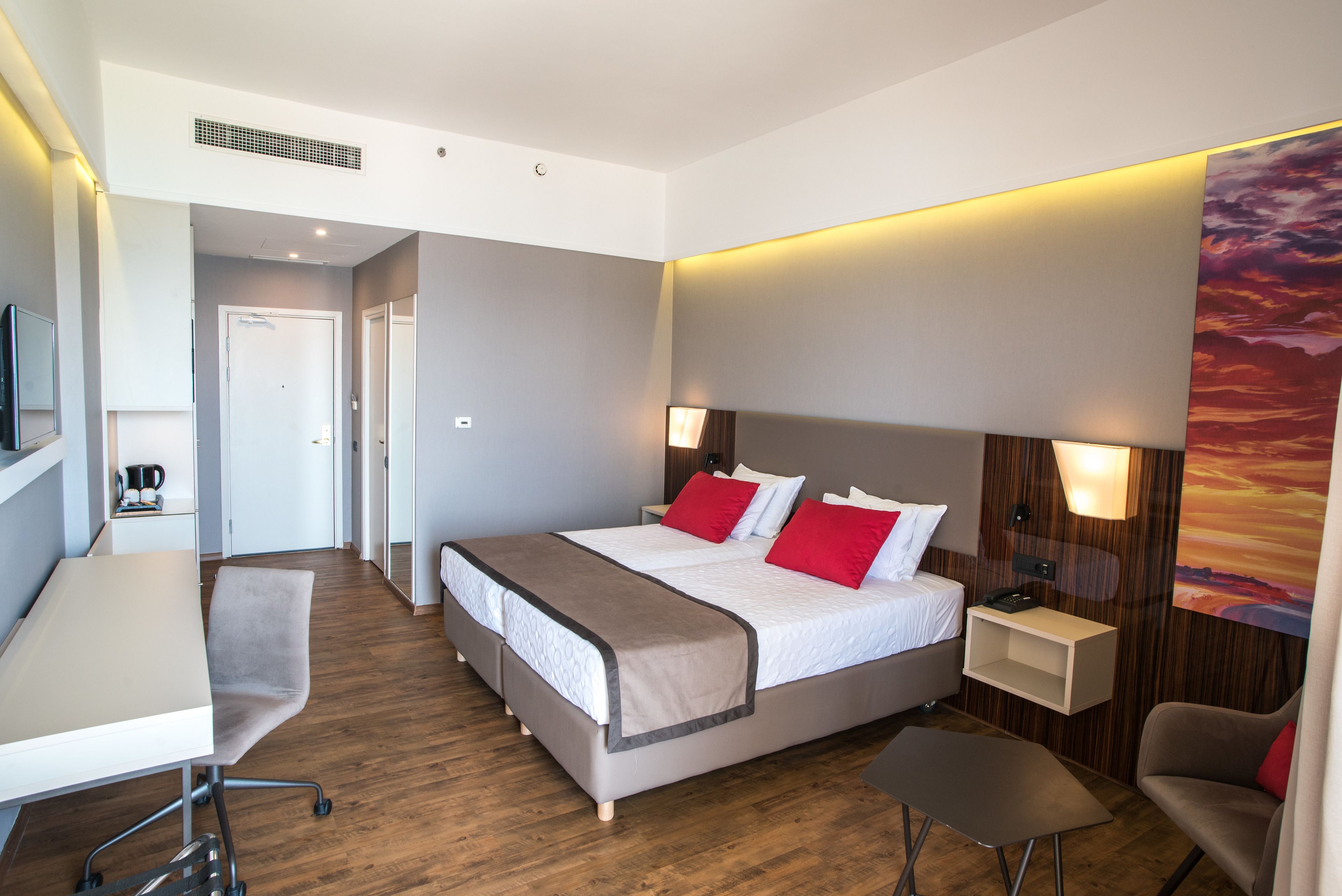 A modern hotel room with a large bed, a desk, and a chair. There is a small table and a nightstand with a lamp.