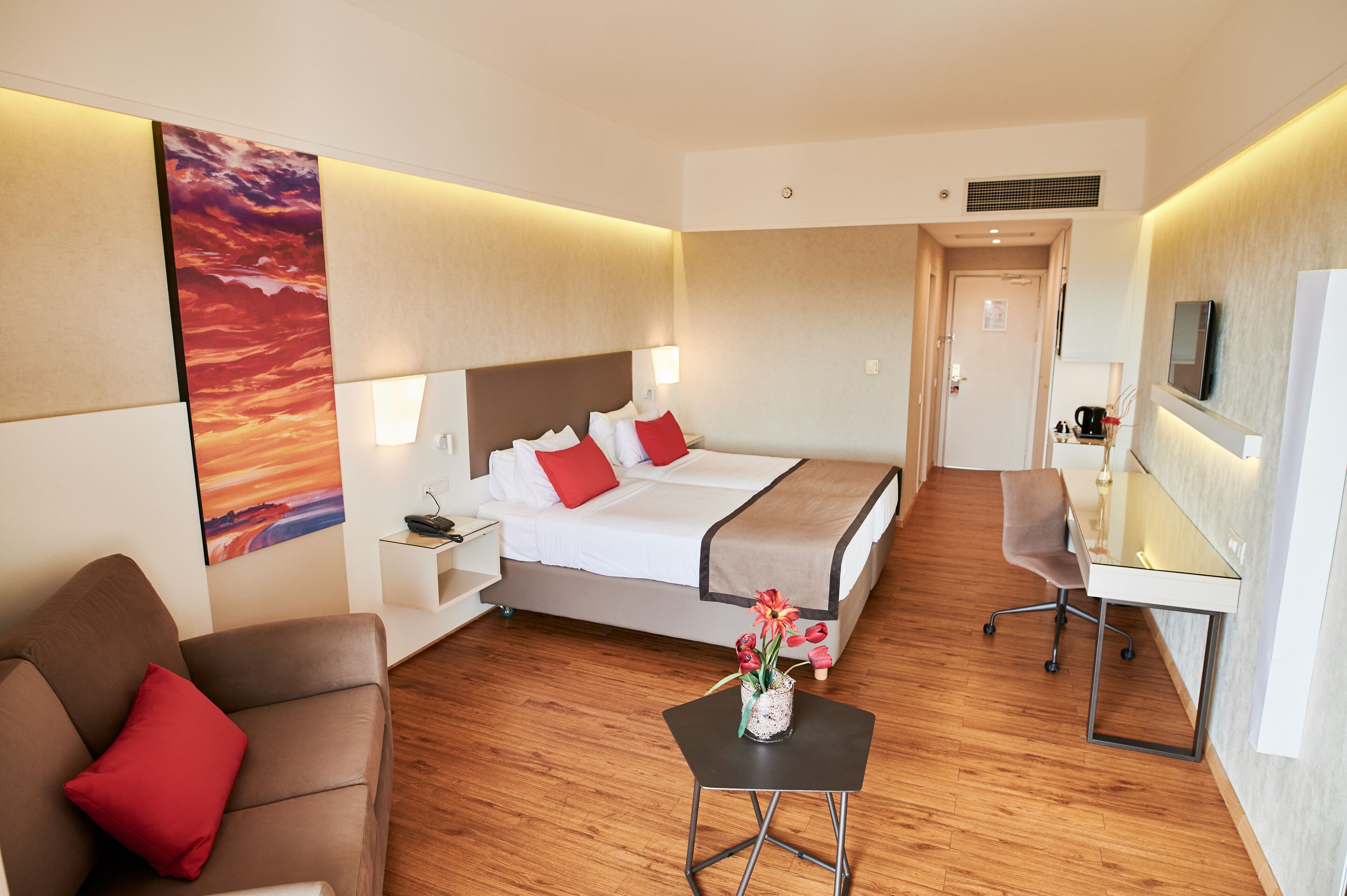 A hotel room with a bed, a desk, a chair, a small table with flowers, a sofa, and a TV on the wall.