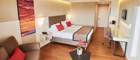 A hotel room with a bed, a desk, a chair, a small table with flowers, a sofa, and a TV on the wall.
