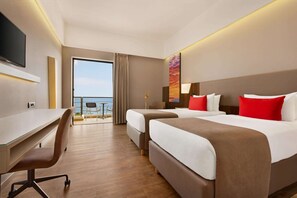 A hotel room with two beds, a desk, a chair, a TV, and a balcony with a view of the sea.