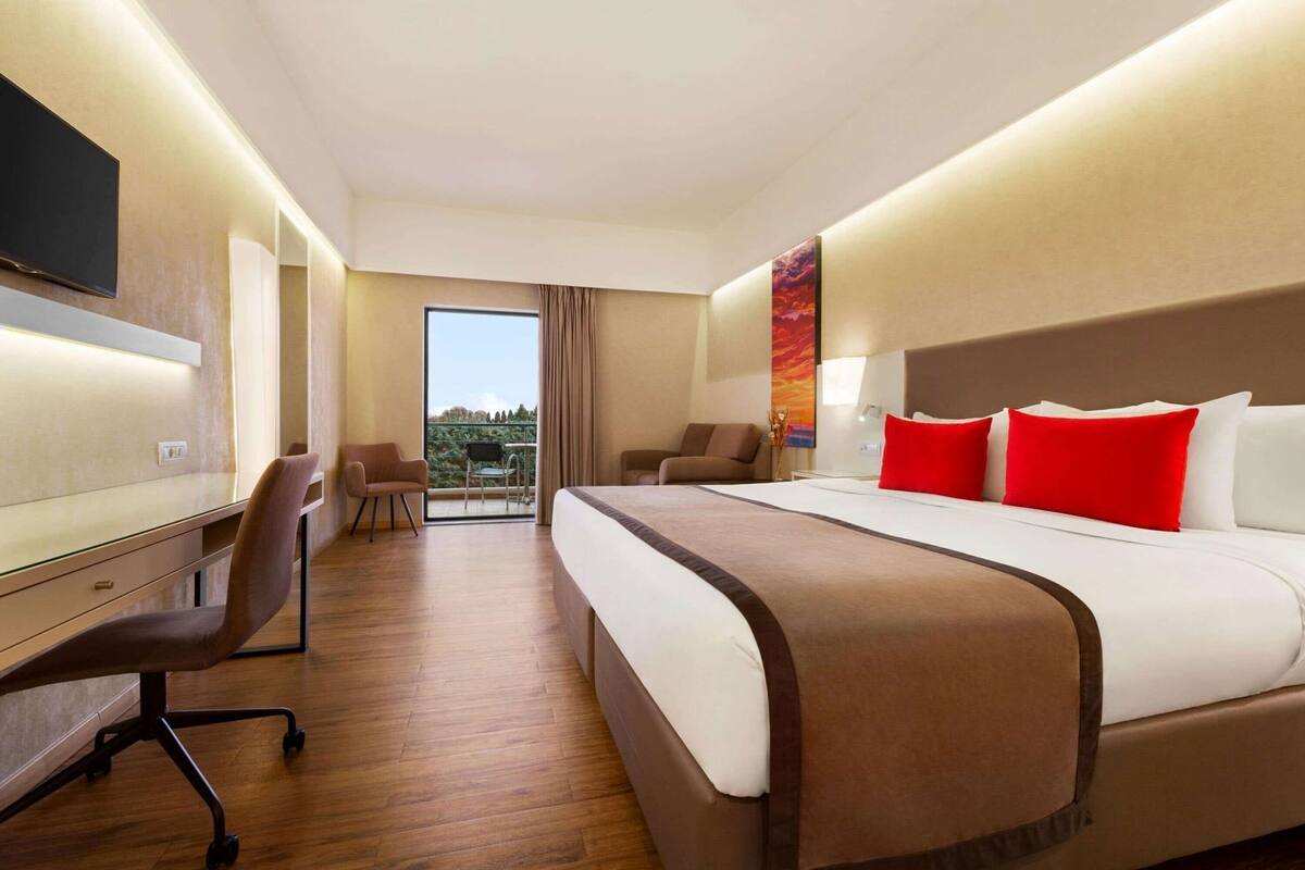 A hotel room with a bed, desk, chairs, and a balcony with a view.