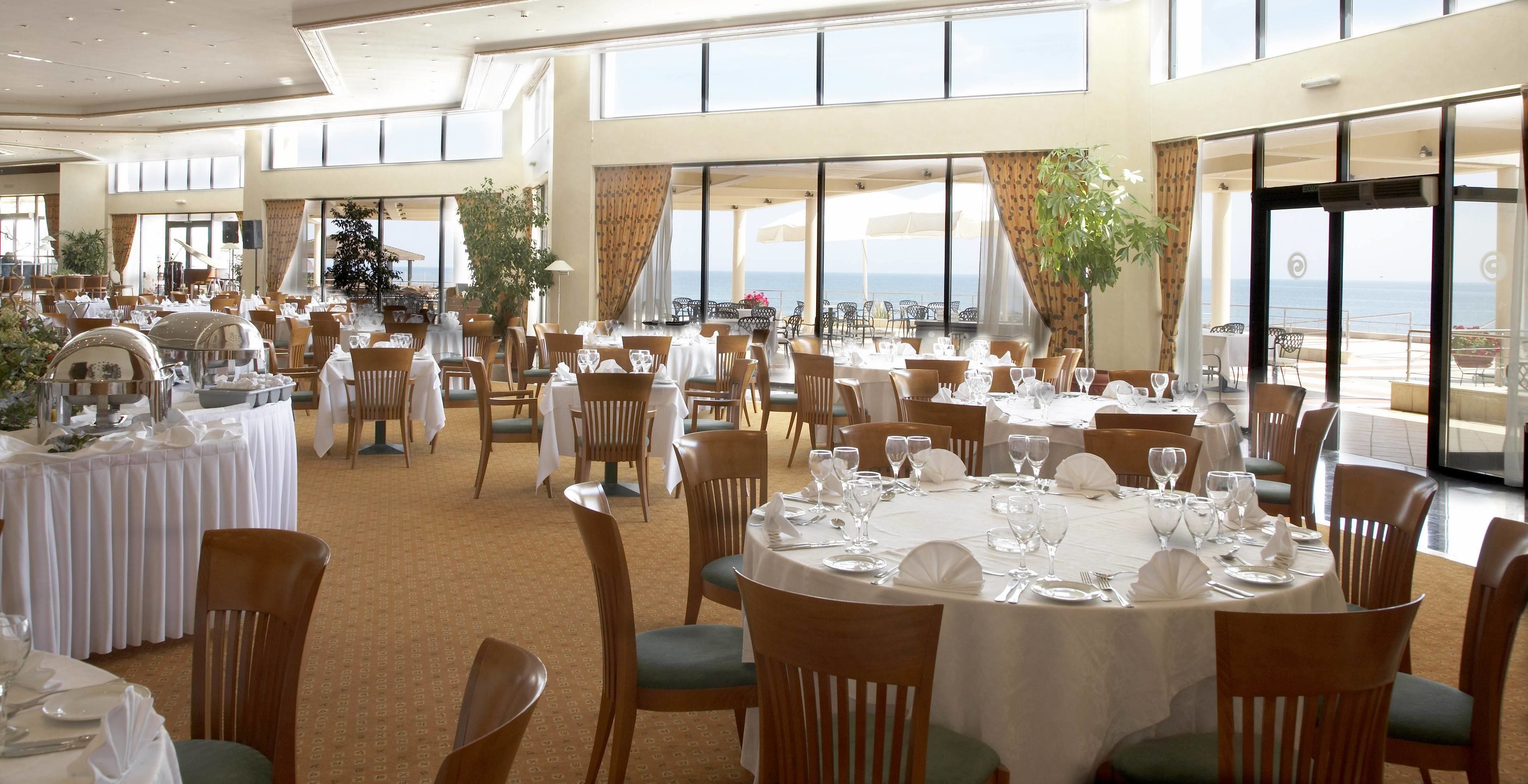 Un vaste espace dédié à la salle à manger, avec des tables rondes disposées pour accueillir de nombreux convives, de grandes fenêtres offrant une vue sur la mer et des plantes en pot qui apportent une touche de verdure.