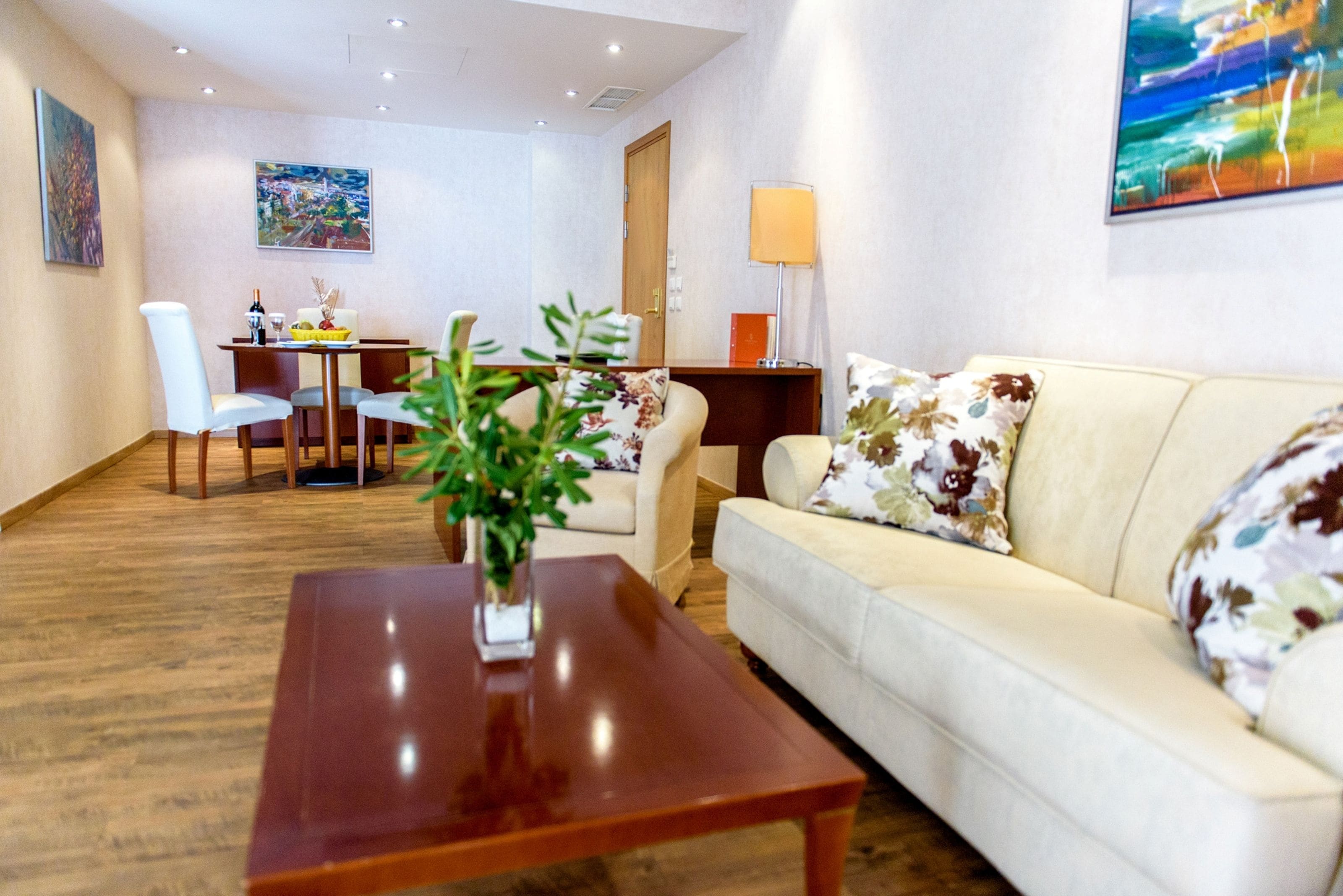 A modern living room with a wooden coffee table, a sofa with floral cushions, a dining area with a table and chairs, and artwork on the walls.