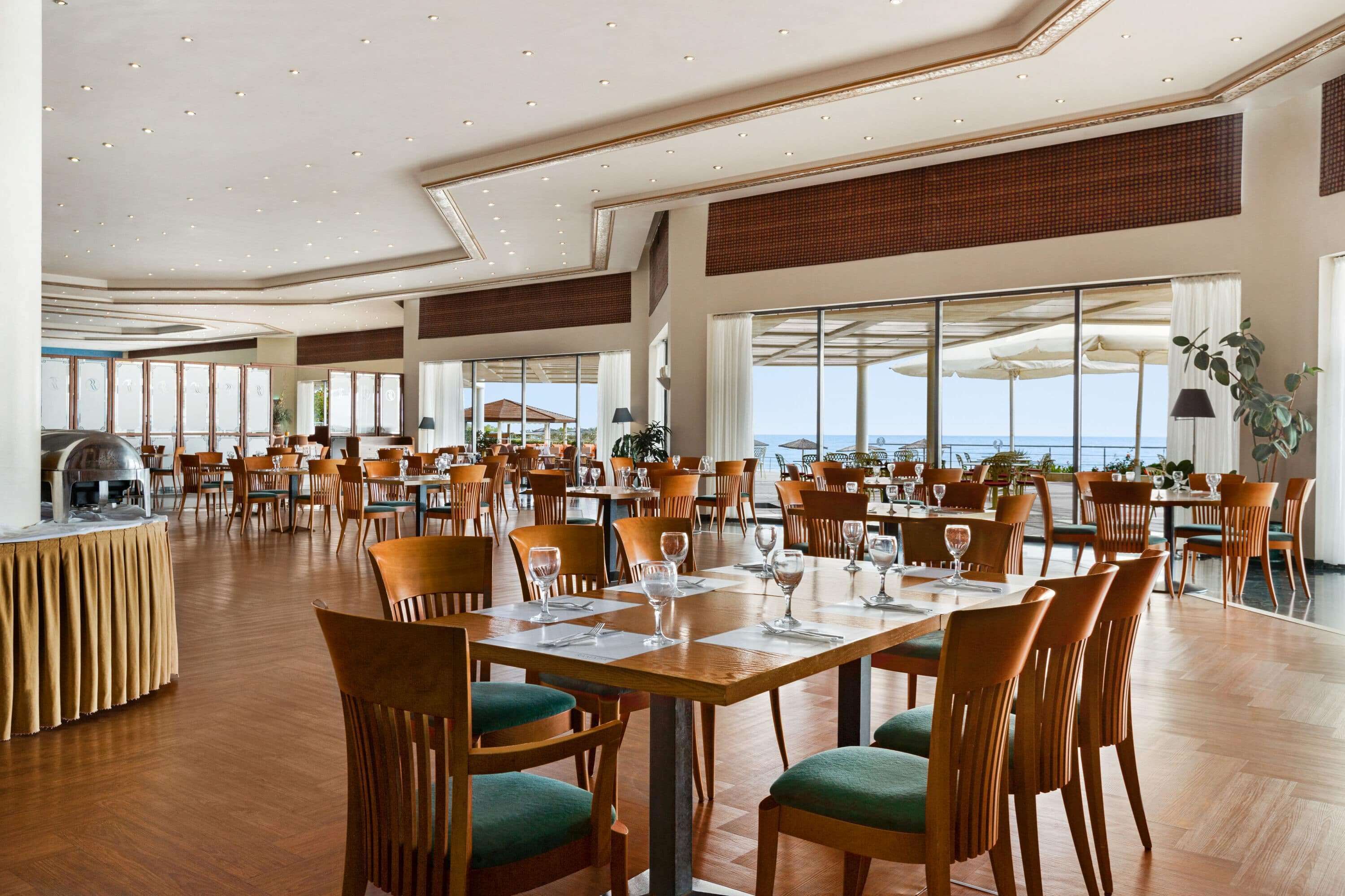 Un comedor amplio con varias mesas y sillas, ventanas grandes que ofrecen vista al mar y un mostrador de buffet con una parrilla.