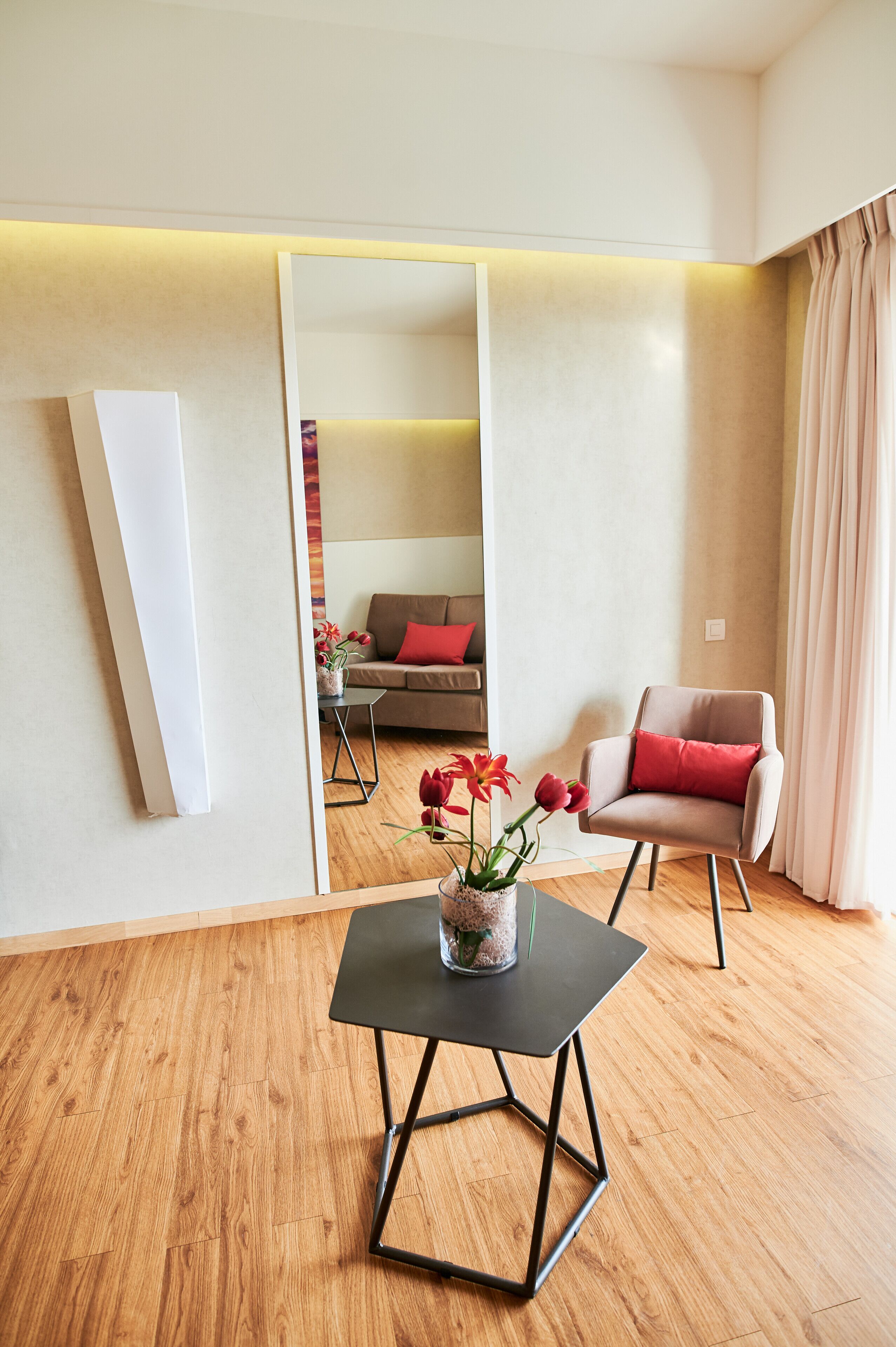 Ein modernes Wohnzimmer mit Holzboden, einem schwarzen Tisch mit Blumenvase, einem beigefarbenen Sessel mit rotem Kissen und einem Sofa mit rotem Kissen.
