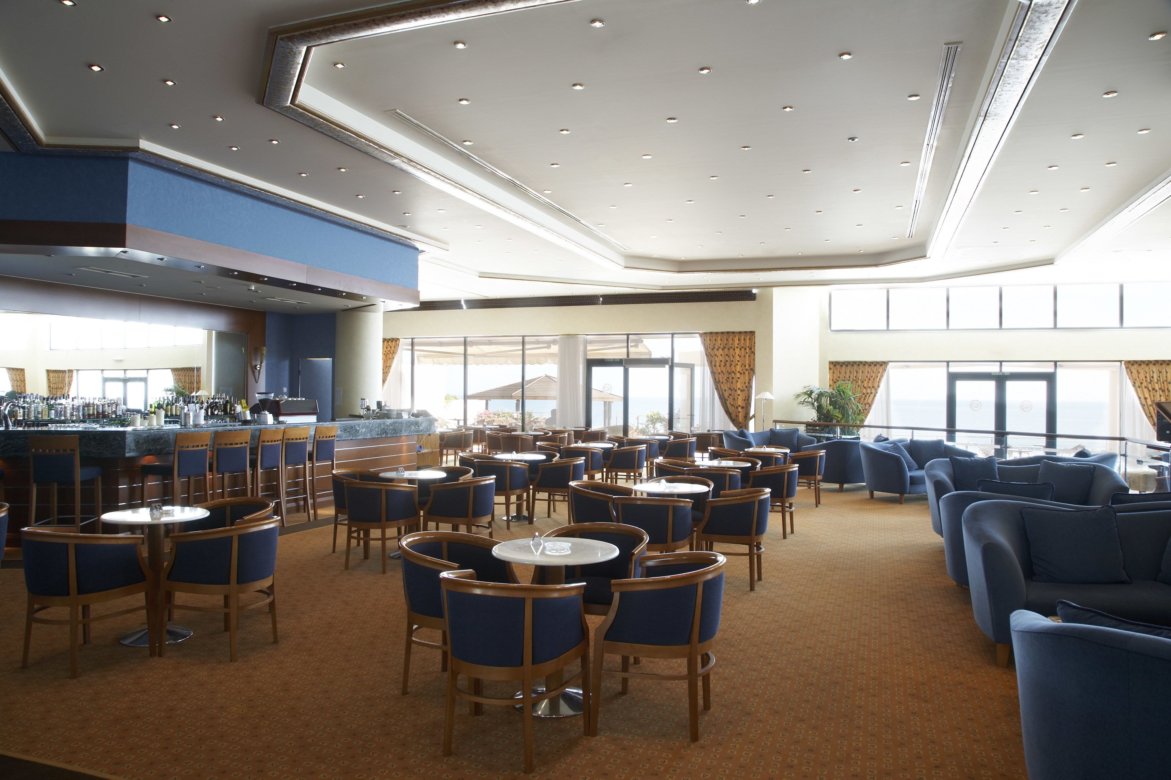 A spacious dining area with multiple tables and chairs, a bar area, and large windows providing natural light.