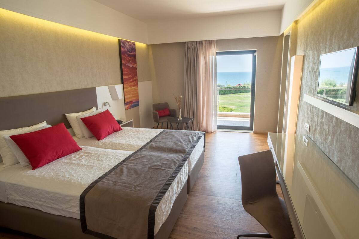 A hotel room with two beds, a chair, a TV, and a view of the sea.