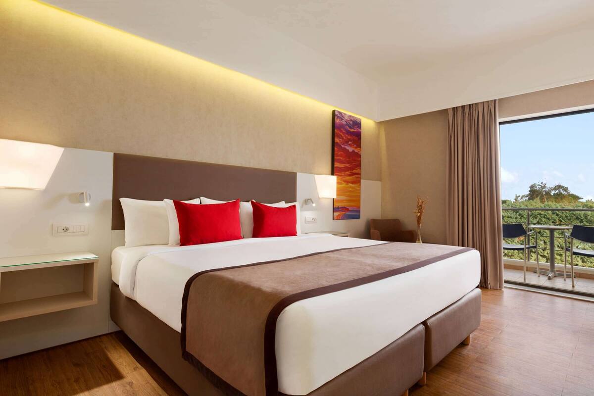 A hotel room with a large bed, bedside tables, a view of the outdoors, and a balcony with chairs.
