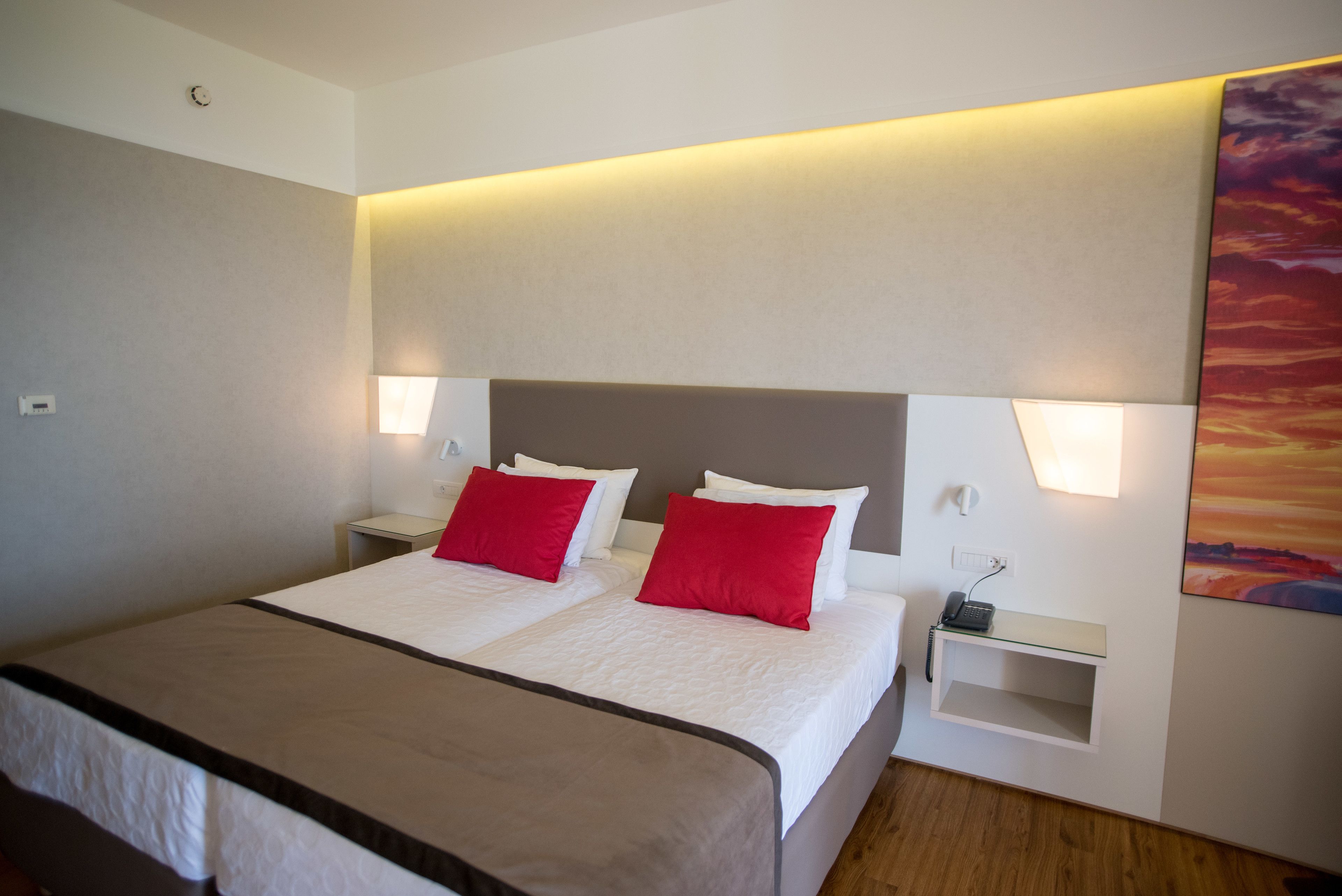A double bed with two red pillows, a bedside table with a phone, and a wall-mounted shelf.