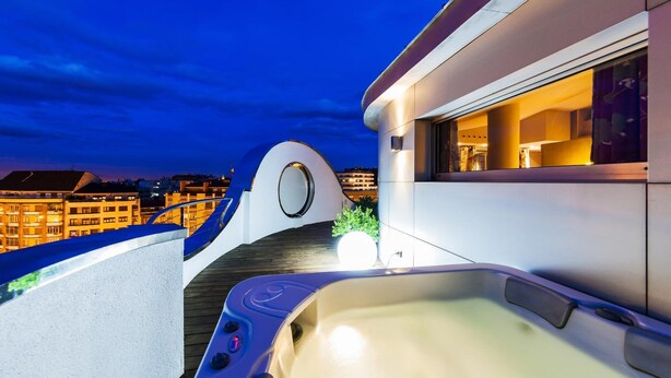 A modern balcony with a hot tub, a wooden deck, and a view of the cityscape at dusk.