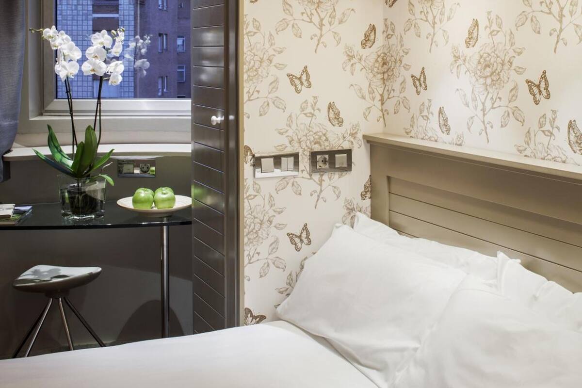 Une chambre à coucher avec un lit, une fenêtre donnant sur des immeubles, une table de chevet avec une plante et des pommes, et un mur recouvert d’un papier peint à motifs floraux.