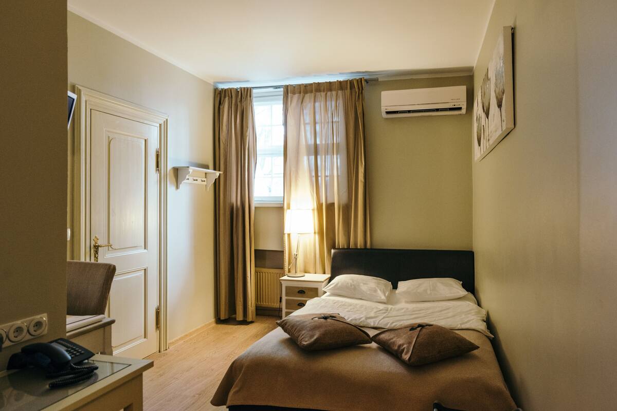 Ein Hotelzimmer mit einem Bett, einem Schreibtisch mit Telefon, einem Nachttisch mit Lampe und einer Klimaanlage an der Wand.