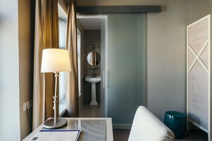 Quarto de hotel com cama, escrivaninha com luminária, janela com cortinas, pia e espelho.
