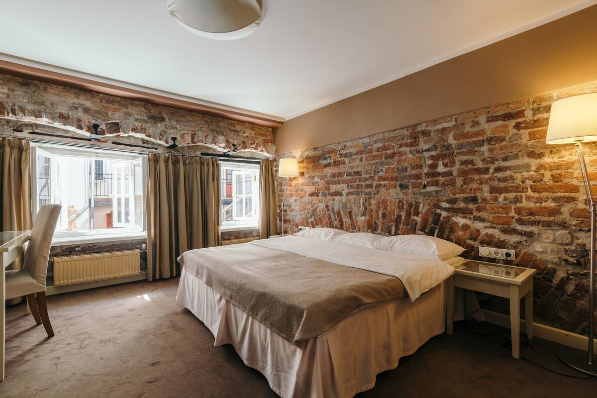 Ein Hotelzimmer mit Backsteinwand, einem großen Bett, einem Schreibtisch mit Lampe und Blick ins Freie.