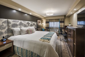 A hotel room with a large bed, a desk with a computer, a TV, and a window with curtains.