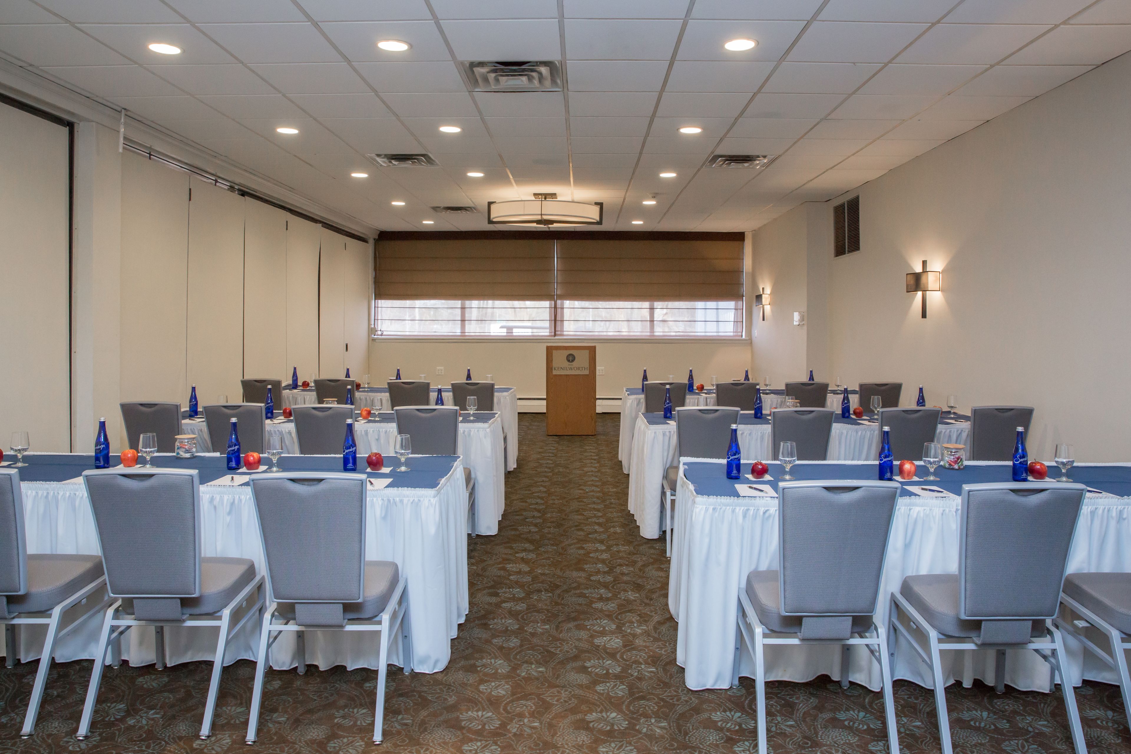 A conference room with rows of tables and chairs, equipped with water bottles and glasses, and a large window providing natural light.