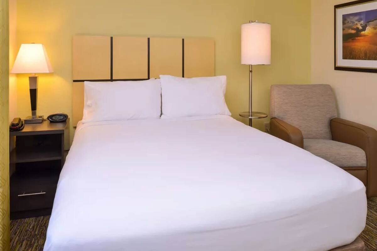 Een net opgemaakt bed met witte lakens, een bedlamp, een klein tafeltje met een telefoon en een afstandsbediening, en een stoel met een kussen.