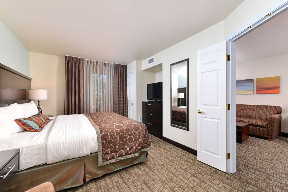 A hotel room with a bed, a desk, a dresser, a TV, a mirror, and a door leading to a living area with a couch.