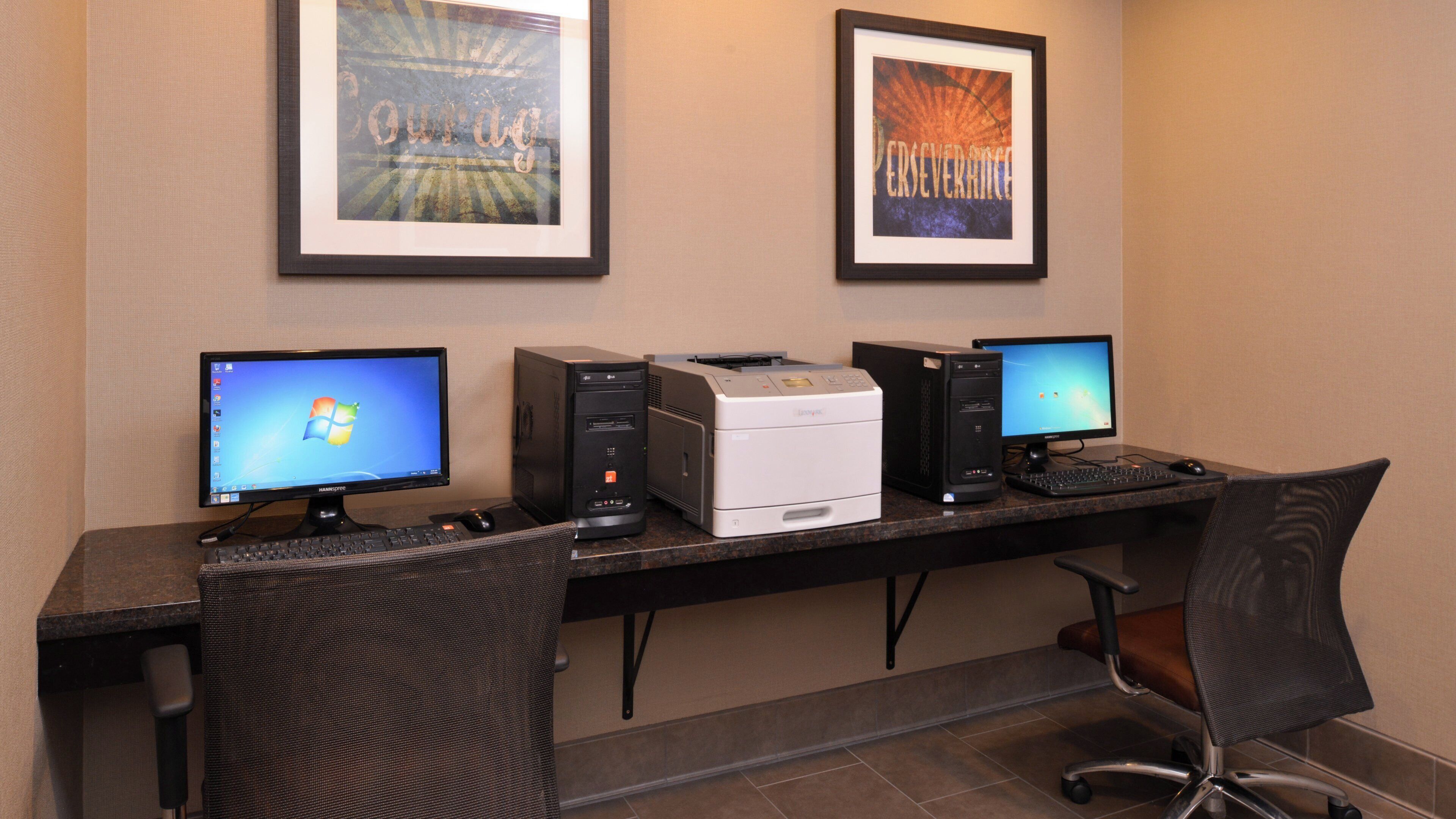 A computer desk with two monitors, a printer, and a keyboard. There are framed pictures on the wall behind the desk.