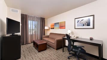 Habitación de hotel con televisor de pantalla plana, sofá marrón con estampado, escritorio con silla, un pequeño cofre de madera y tres cuadros en la pared.