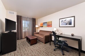 A hotel room with a flat-screen TV, a sofa, a desk with a chair, a small table, and artwork on the wall.