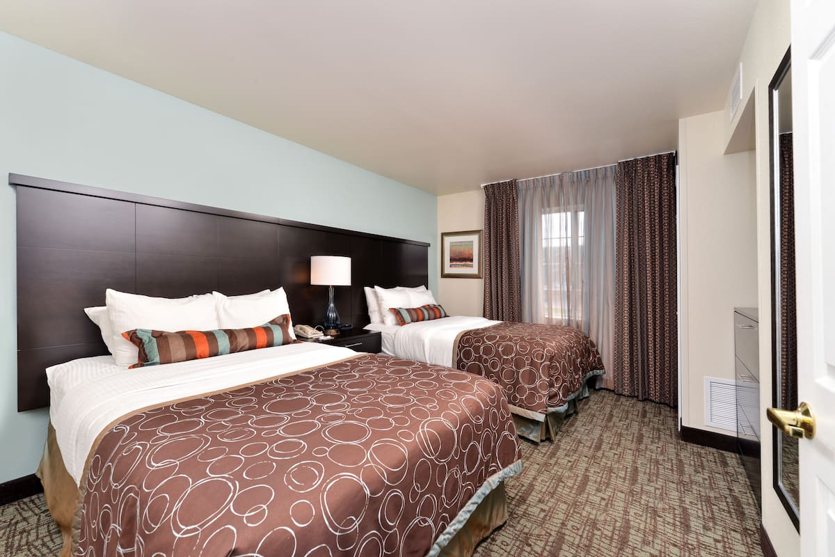 A hotel room with two beds, a dark headboard, a nightstand with a lamp, and a window with curtains.