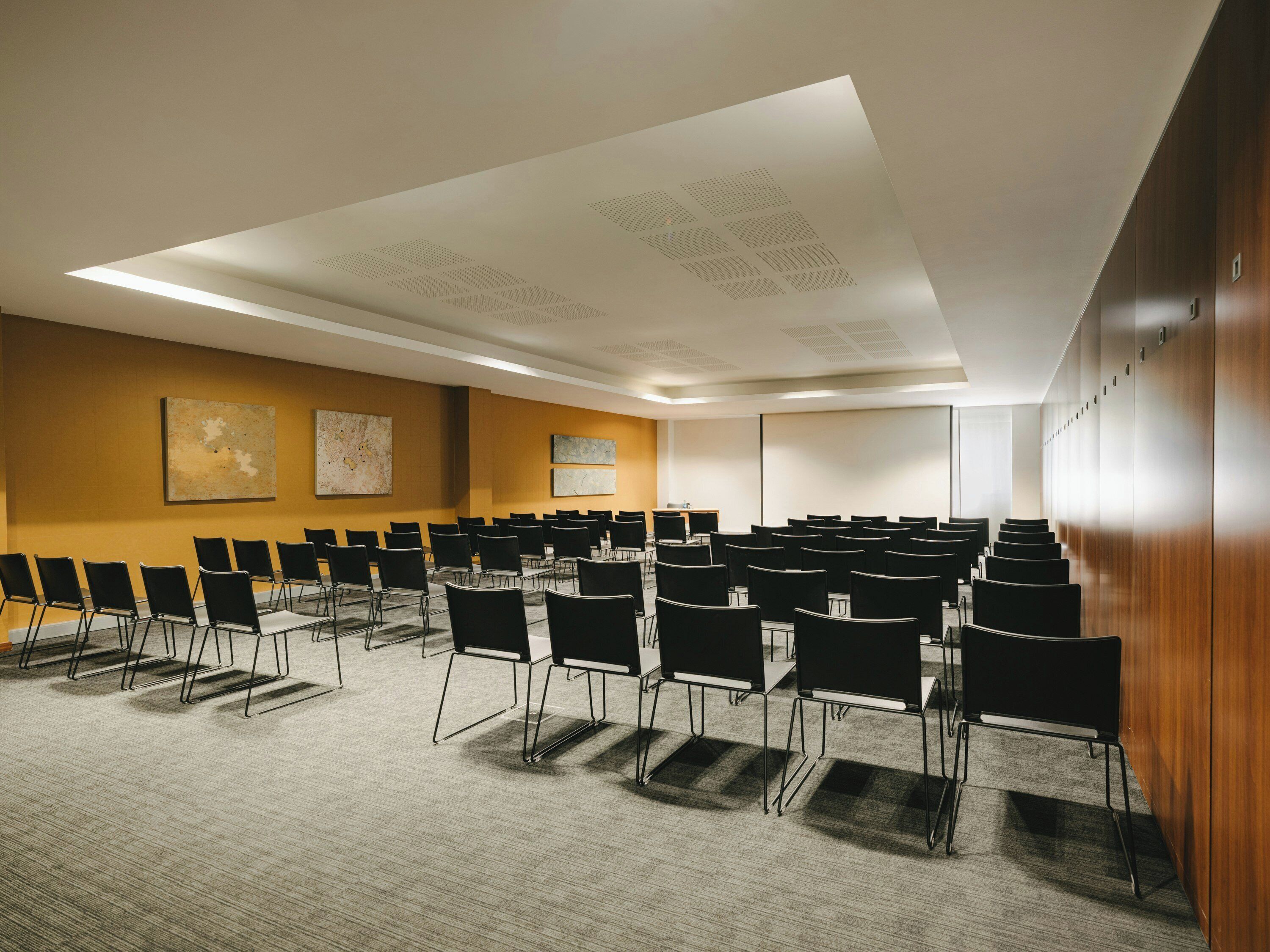 Un salón de conferencias con filas de sillas, una pantalla de proyector y un cuadro abstracto grande en la pared.