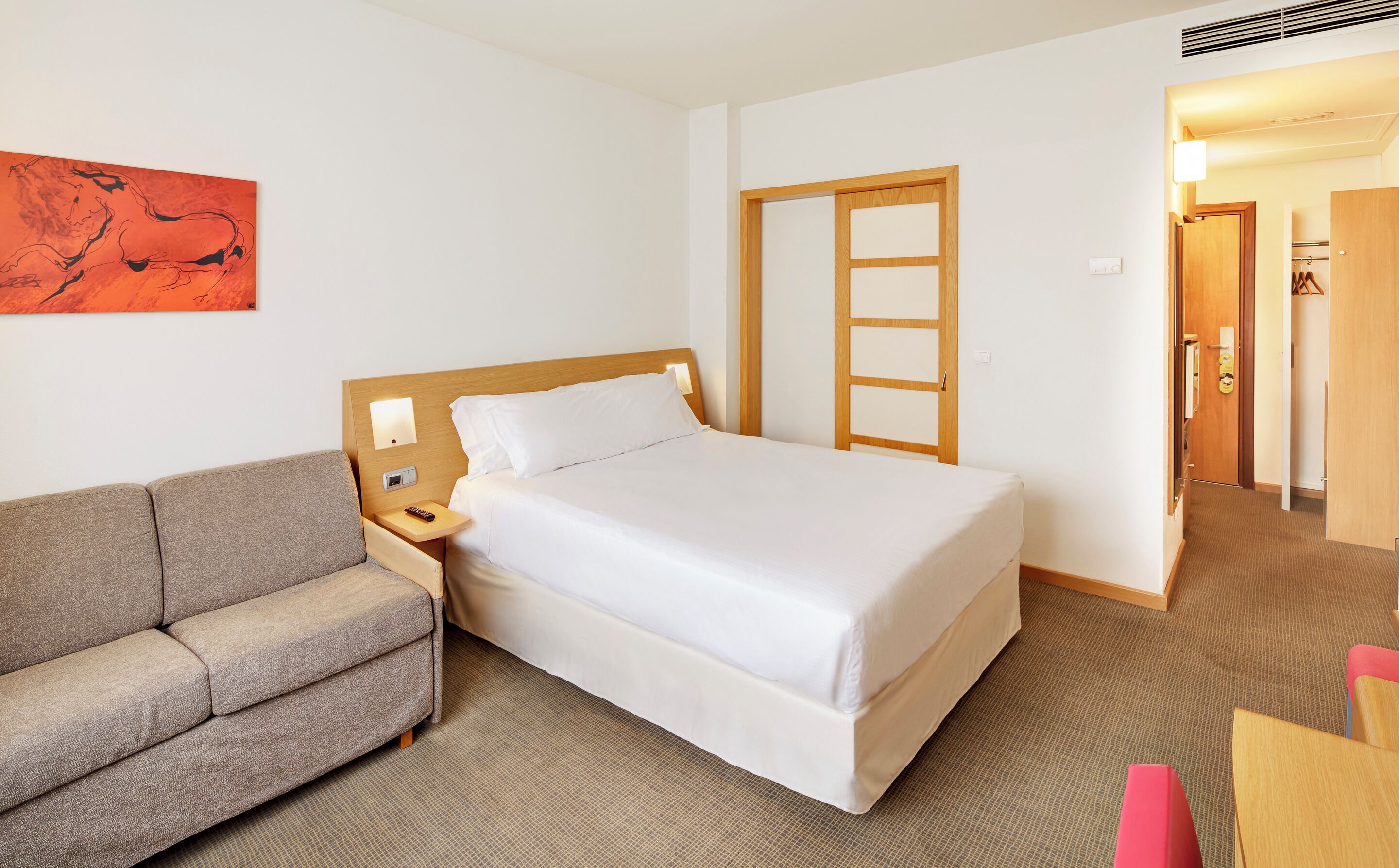 Quarto de hotel com cama, sofá, mesa de cabeceira, controle remoto e um quadro na parede.
