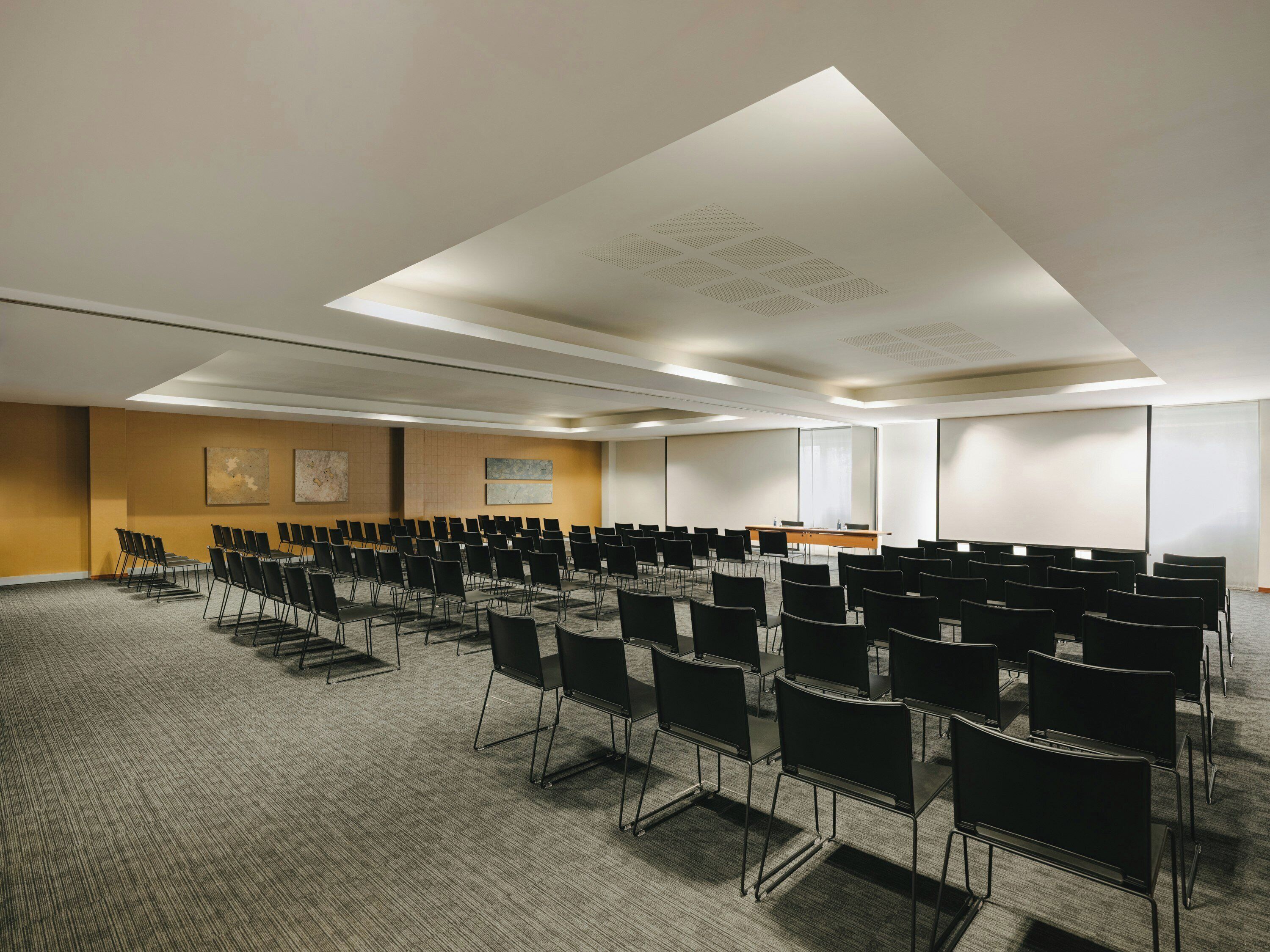 Uma sala de conferências com fileiras de cadeiras, uma mesa e uma tela de projeção.