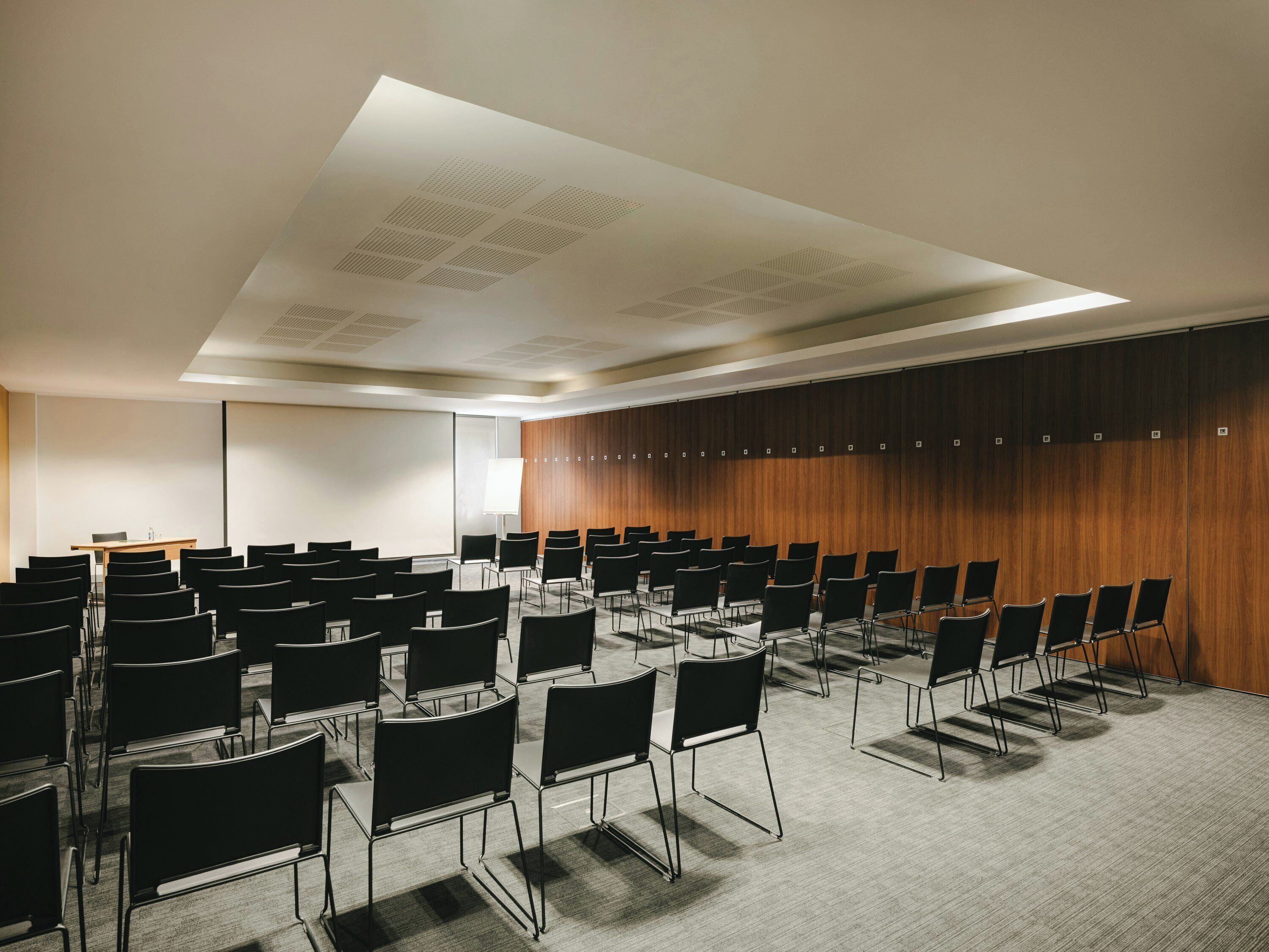 Una sala de conferencias con filas de sillas, una pantalla de proyector y un panel de pared de madera.