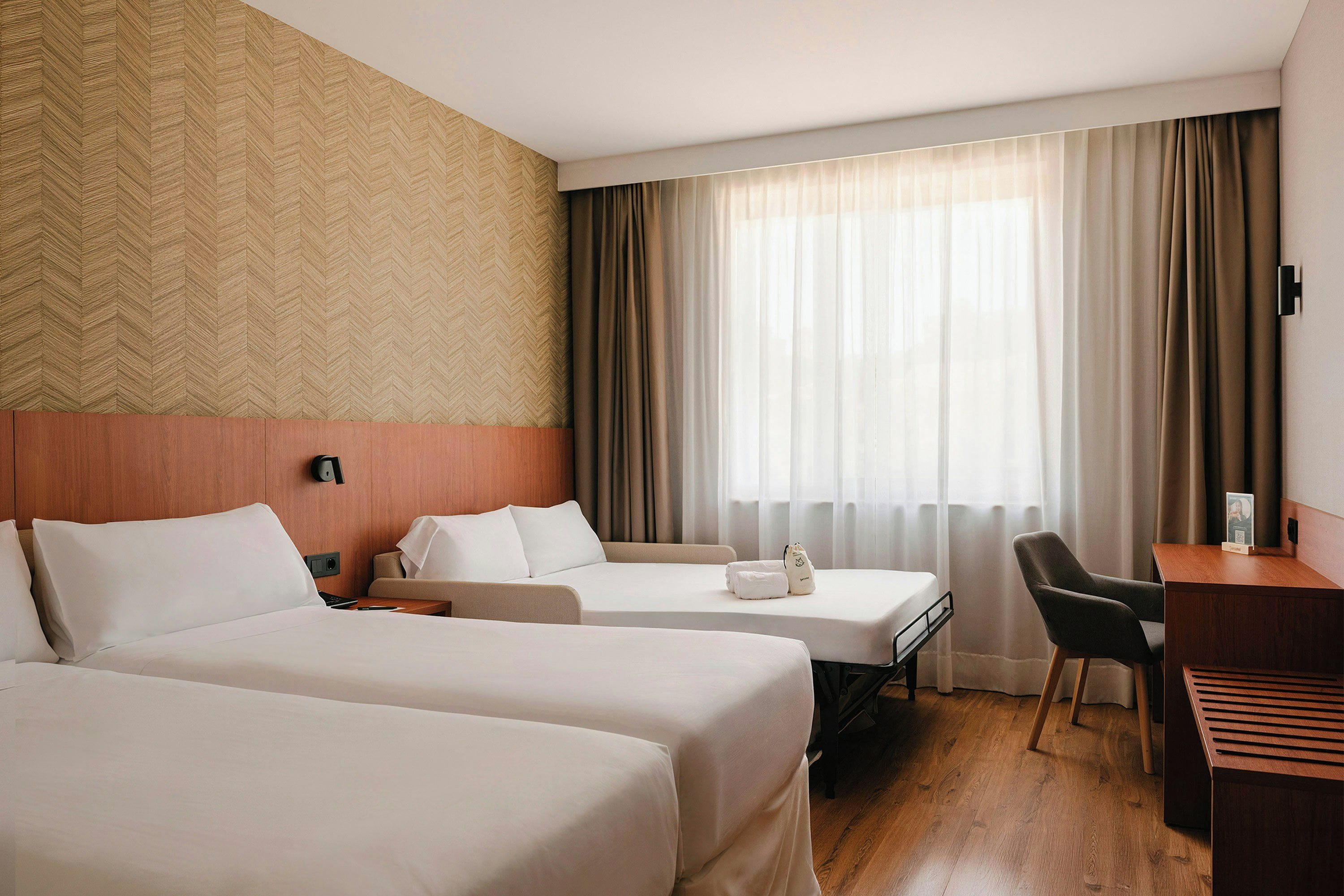 Habitación de hotel con dos camas, un escritorio, una silla y un amplio ventanal con cortinas.