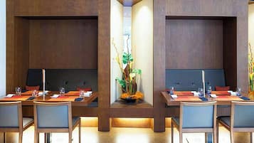 Un comedor moderno con mesas y sillas de madera, una planta decorativa en el centro y un espacio de estar empotrado.