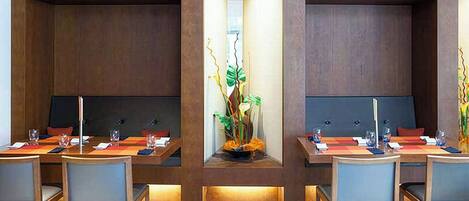 A modern dining area with wooden tables and chairs, a central decorative plant, and a recessed seating area.