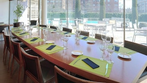 A long dining table set with chairs, glassware, and plates, with a view of buildings and trees through large windows.